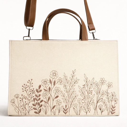 Beige tote bag with floral embroidery and brown leather straps on a white background