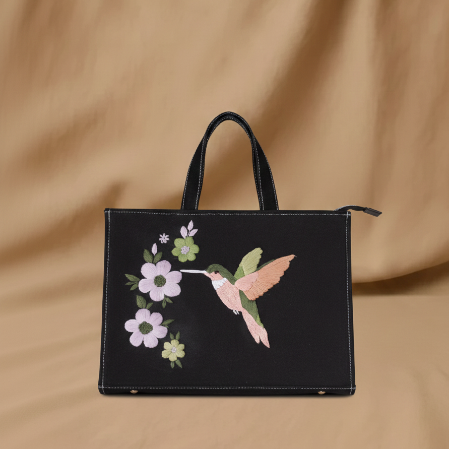 Black tote bag with embroidered hummingbird and flowers on a beige background